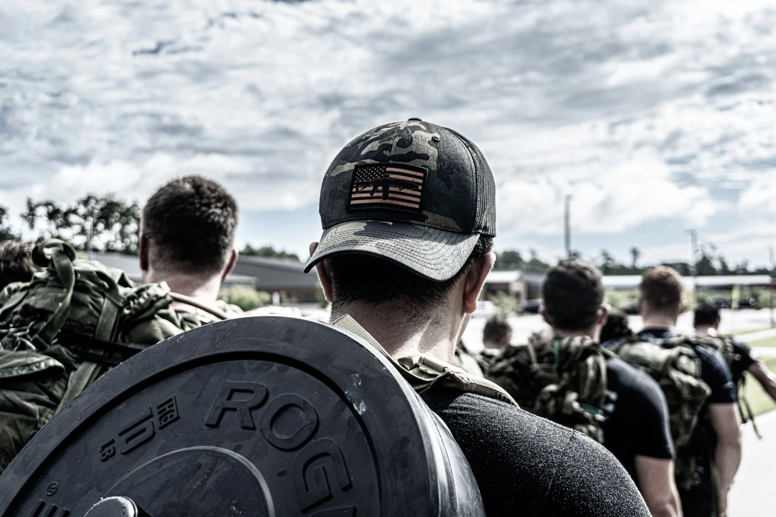 Seen from behind, a competitor shoulders a Rogue weight plate while a line of ruckers heads up the road