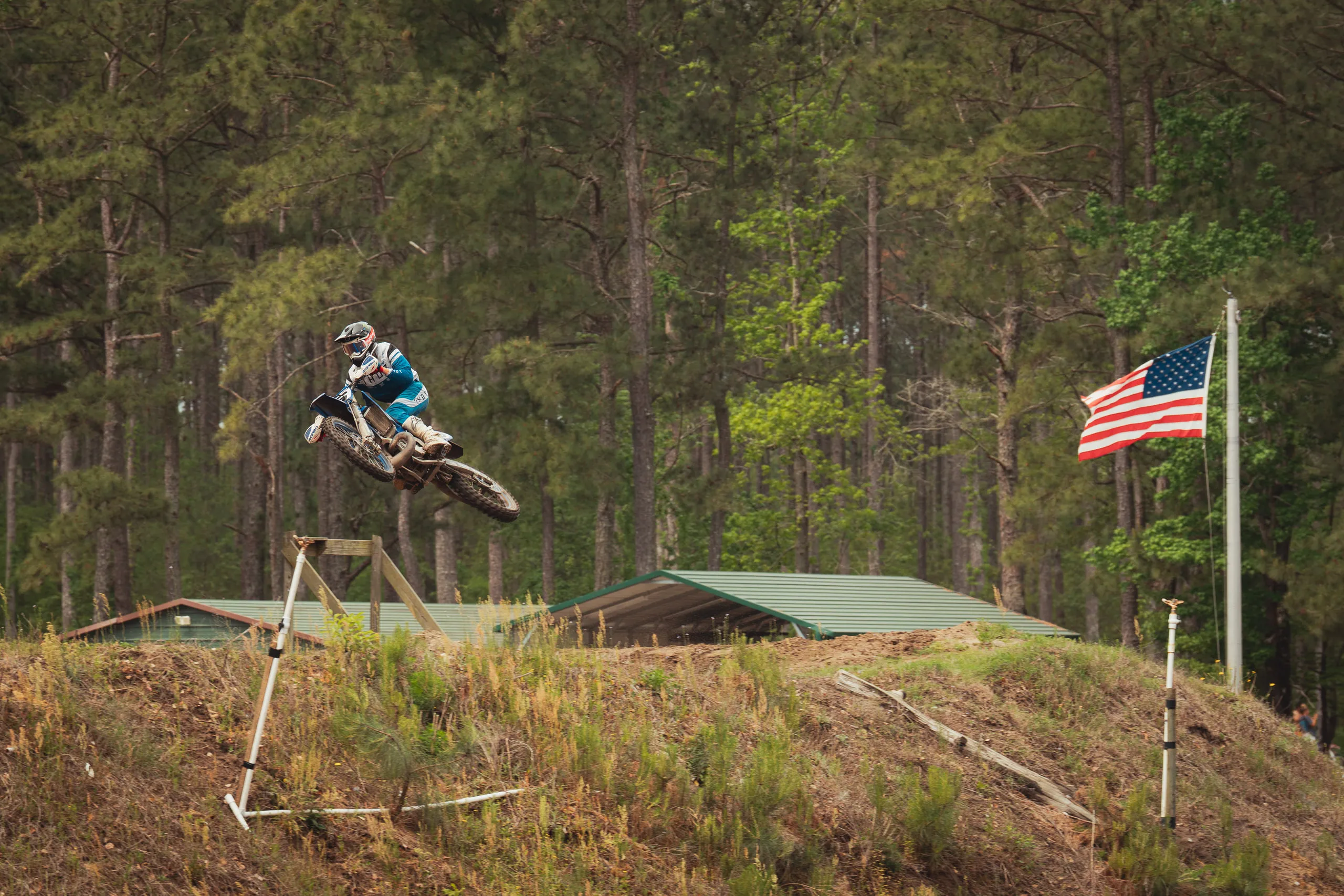 Rider launches off a tabletop jump past an American flag with pine woods behind